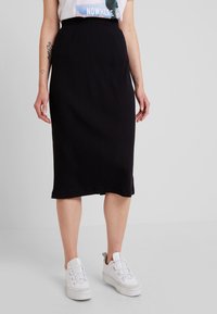 Jupe midi noire à côtes avec taille élastique, associée à des baskets blanches. La jupe présente une coupe droite et une finition à l'ourlet.