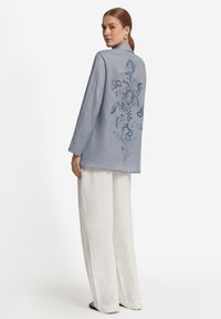 Ljusblå linnéjacka med långärmar, prydd med blommig broderi på baksidan. Matchad med lösa vita byxor, minimalistisk design.