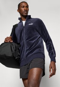 Tume sinine sametist kapuutsiga jakk, millel on kõrge kaelus, koos mustade lühikestega. Tumeda musta spordikott hoitakse ühes käes. Valge logo rinnal.