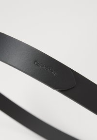 Zwarte leren riem met een gladde textuur, voorzien van een geperst "Calvin Klein" logo op de band. Minimalistisch ontwerp zonder extra versieringen.