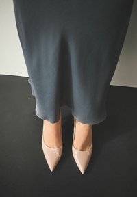 Beige lakleren pumps met een spitse neus en een strak ontwerp, gedragen bij een donkergrijze, vloeiende jurk die zichtbaar is aan de zoom boven de schoenen.