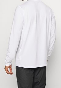 Man som bär en enkel vit långärmad skjorta och svarta byxor, sedd bakifrån, stående mot en enkel ljus bakgrund.