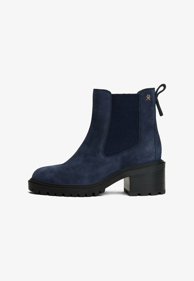 Bottines navales en daim avec panneaux latéraux élastiques, un talon texturé, une semelle en caoutchouc et une tirette, mettant en avant un accent de logo subtil.