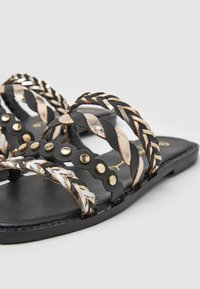 Sandales plates noires avec des brides tressées en noir, or et argent métallique. Comportent des clous dorés le long des brides et une semelle lisse et texturée.
