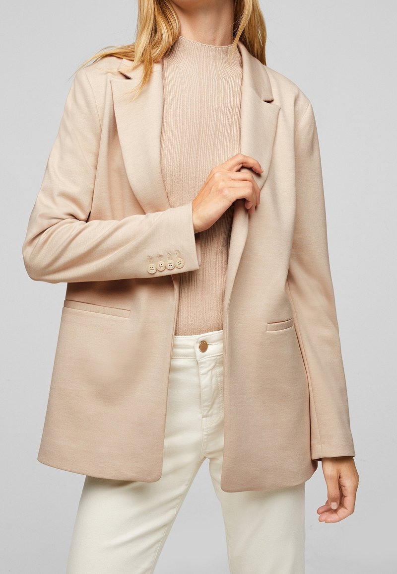 Světlobéžový blazer vyrobený z měkkého materiálu; má klopový límec, dvě přední kapsy a manžety na knoflíky, doladěný s béžovým žebrovaným topem.
