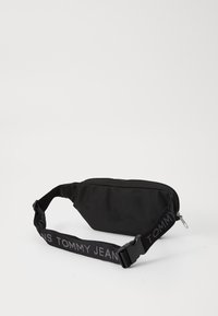 Zwarte stoffen heuptas met een ritsvak, voorzien van een ingenaaide band met de tekst "TOMMY JEANS" in grijs. Compacte en rechthoekige vorm.
