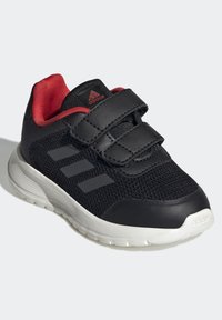 Černá sportovní bota s síťovaným svrškem, červenými akcenty, suchým zipem, bílou podrážkou a třemi šedými pruhy po straně. Lehká konstrukce.