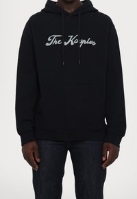 Muž v černé mikině s kapucí a přední kapsou, s bílým kurzivním nápisem "The Kooples", v kombinaci s tmavě modrými džíny, stojící na jednobarevném pozadí.