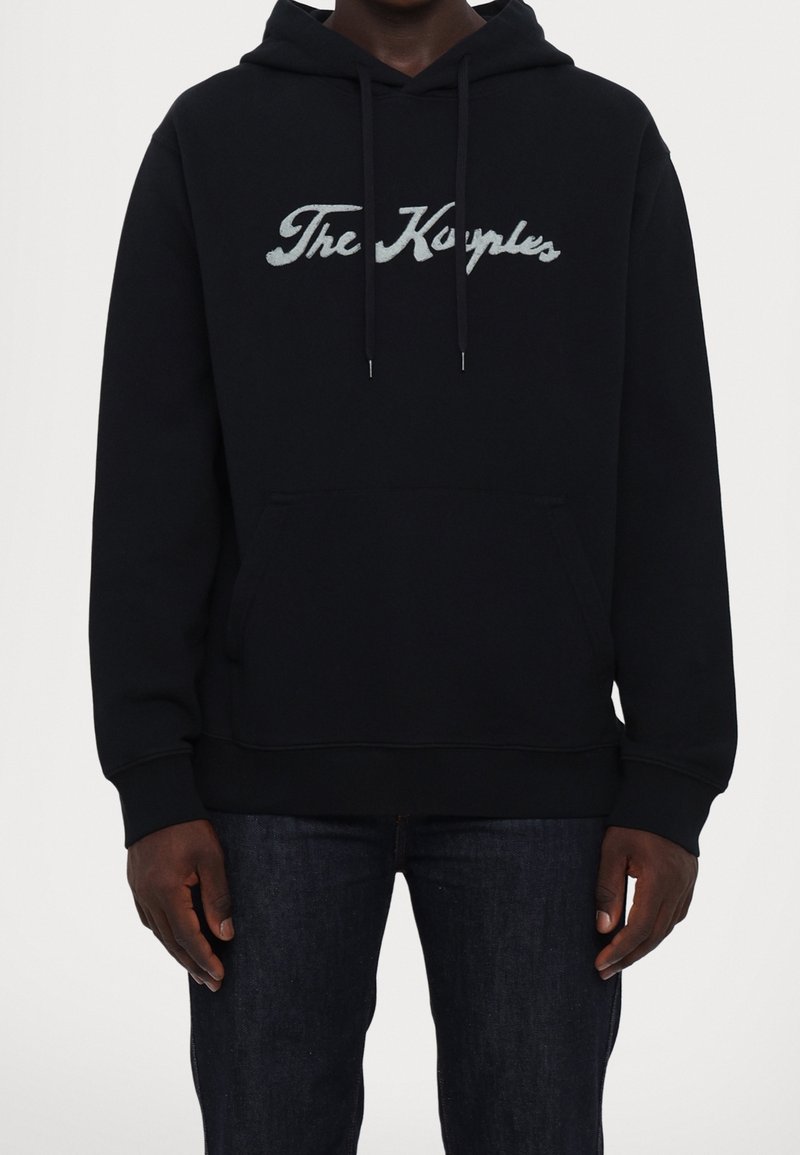 Muž v černé mikině s kapucí a přední kapsou, s bílým kurzivním nápisem "The Kooples", v kombinaci s tmavě modrými džíny, stojící na jednobarevném pozadí.