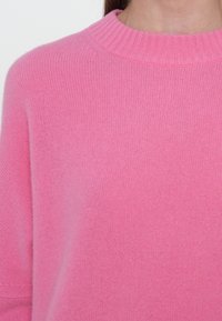 Femme portant un pull en tricot rose pâle avec un col rond côtelé, visible des épaules jusqu'au milieu de la poitrine, sur un fond blanc.