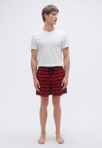 Κόκκινα και μαύρα καρό σορτς με ελαστική μέση και κορδόνι, συνδυασμένα με ένα απλό λευκό μπλουζάκι. Μοντέλο όρθιο με γυμνά πόδια.