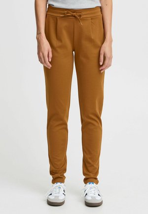 Hnědé joggingové kalhoty vyrobené z měkké tkaniny, s elastickým pasem na šňůrku, bočními skladbami a kotníkovými manžetami. V kombinaci s bílými teniskami.