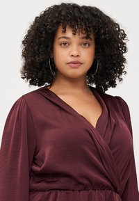 Woman with curly hair wears a deep burgundy dress with a wrap design, showcasing her natural beauty and confidence.