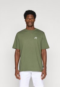 Olīvu zaļa kokvilnas T-kreklu ar īsām piedurknēm, apaļu kakla izgriezumu un baltu logo kreisajā krūtī. Taisna apakšmala, brīva piegriezuma.