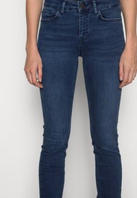 Jeans ajustados hechos de denim azul oscuro, que cuentan con cinco bolsillos, cinturilla, y un ligero desgastado en la parte delantera, con cierre de botón.