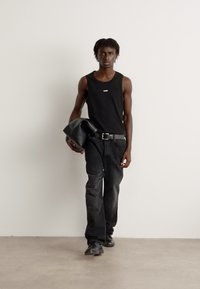Top negru fără mâneci cu un mic logo, blugi negri din denim și o jachetă neagră ținută într-o mână. Design elegant și modern, cu linii simple.