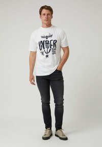 Camiseta blanca de algodón con un gráfico azul marino que incluye un ancla y texto, combinada con pantalones oscuros y zapatillas beige. Diseño simple y casual.