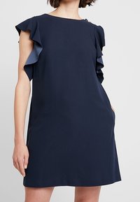 Vrouw in een marineblauwe mouwloze jurk met gerimpelde schouderdetails tegen een effen lichte achtergrond.