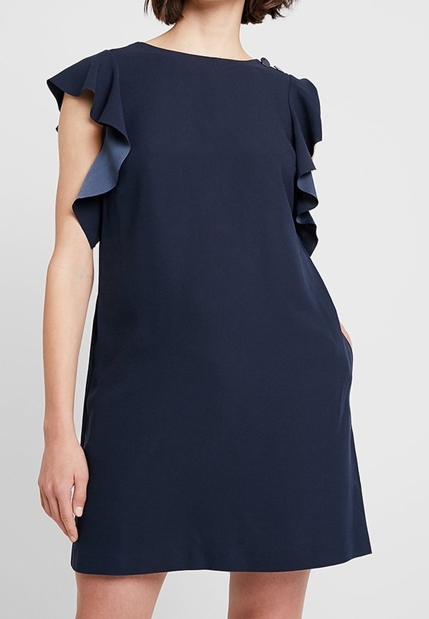 Vrouw in een marineblauwe mouwloze jurk met gerimpelde schouderdetails tegen een effen lichte achtergrond.