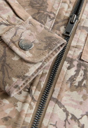 Kamuolių rašto audinys smėlio ir rožinės spalvų atspalviais, su užtrauktuko uždarymu ir metalinio mygtuko detale. Tekstūruotas paviršius.