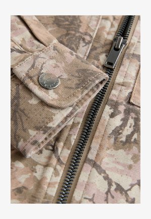 Maastokuvioinen kangas, jossa on beigejä ja vaaleanpunaisia sävyjä, vetoketjusuljenta ja metallinen nappidetalji. Pinta on teksturoitu.