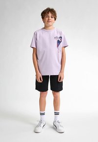 Lilaanin värinen t-paita, jossa vasemmalla puolella oleva painettu flamingokuosi, yhdistettynä mustiin cargo-shortseihin ja valkoisiin lenkkareihin, joissa mustia yksityiskohtia.