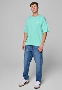 Miętowa, oversize'owa koszulka z kieszonką na piersi, na której widnieje napis "WILLIAMS" w czerni. Uzupełniona niebieskimi dżinsami i białymi trampkami z brązowymi akcentami.