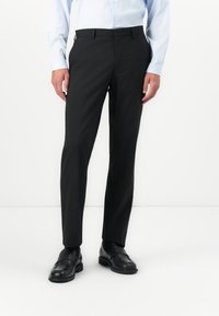 Mann iført svarte dressbukser, svarte skinn loafers og en lyseblå skjorte med lange ermer som står på et hvitt gulv.