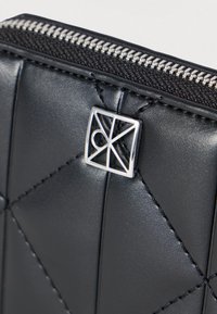 Cartera de cuero negro acolchada con una placa de logo plateada y una cremallera en contraste. Textura suave con detalles de costuras precisas.