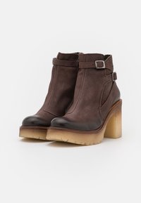 Botas de cuero marrón hasta el tobillo con punta negra, tacón grueso beige y hebillas plateadas en los lados. Superficie texturizada y bordes suaves.