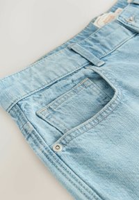 Un jean en denim bleu clair présente une poche avant, des détails de couture et une ceinture avec une boucle. Texture lisse avec un aspect légèrement usé.