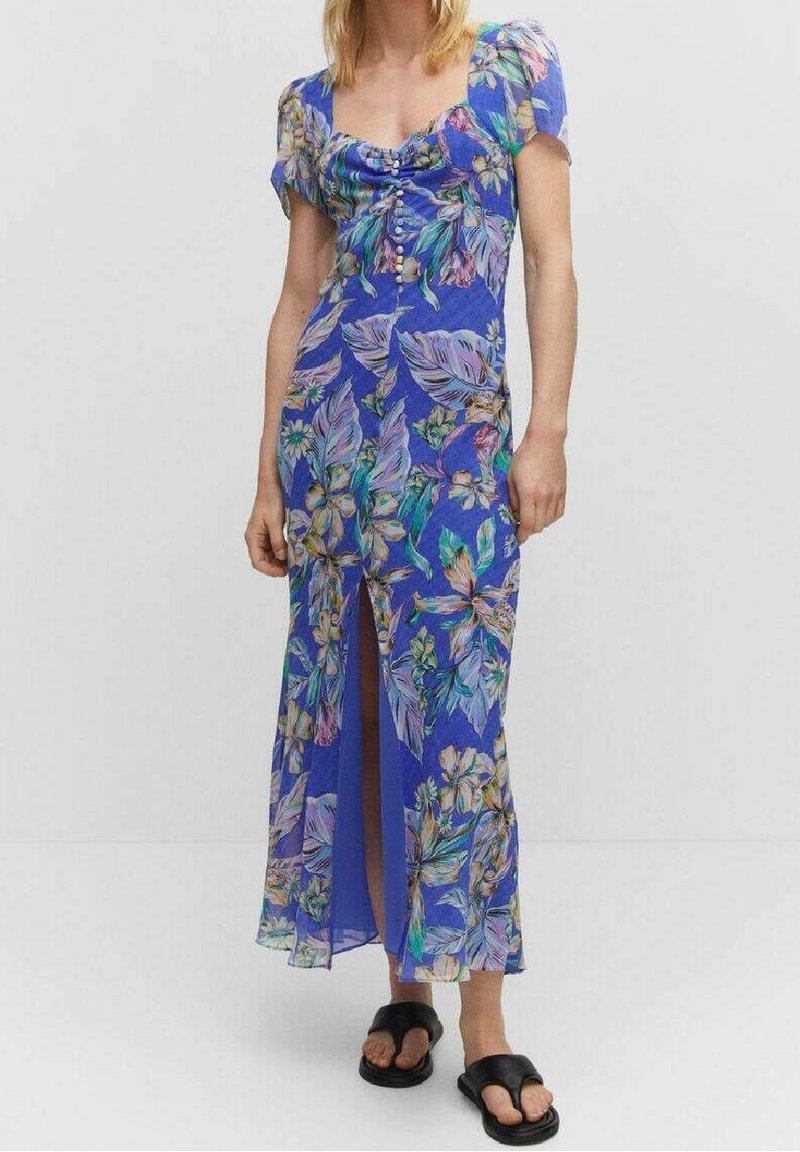 Femme portant une robe midi bleue à fleurs, manches courtes, fente devant et boutons, associée à des sandales noires sans brides.