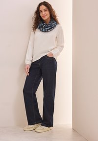 Hvid langærmet skjorte, mørkeblå denim bukser med brede ben, mønstret blå og sort uendelighedsscarf, beige sneakers, står foran en lys væg.