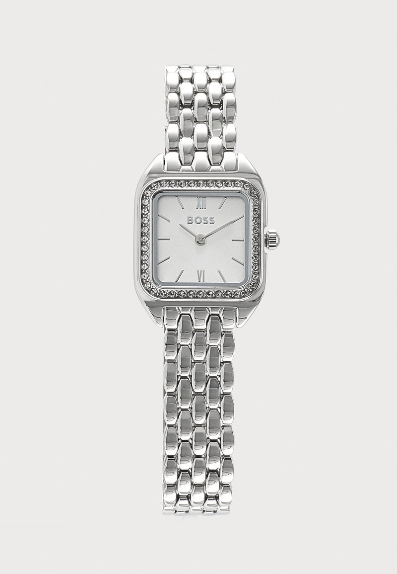 Montre-bracelet carrée argentée avec lunette ornée de cristaux, cadran blanc, chiffres romains et bracelet métallique articulé.