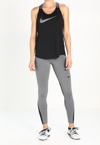 Nainen, joka pukeutuu mustaan Nike-työhön, harmaisiin Nike-leggings-housuihin mustilla raidoilla ja valkoisiin urheilukenkiin, seisoo valkoista taustaa vasten.