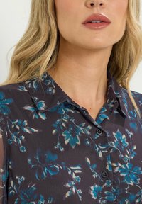 Blusa floreale in tessuto scuro con motivi di fiori blu e crema, colletto classico e bottoni neri davanti.