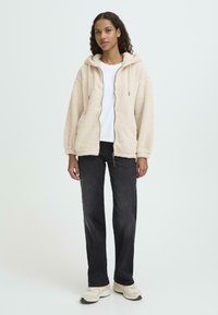 Bež fliisist kapuutsiga jakke, mis on lukuga ja nööridega, kantud valge t-särgi peal ning kokku pandud mustade sirgete pükste ja heledate tossudega.