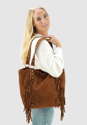 Blonde woman in white sweater holds large brown suede fringe handbag over shoulder, smiling slightly, against plain background.