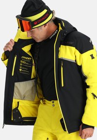 Schwarze und gelbe Spyder-Skijacke, ausgestattet mit wasserdichtem Material, seitlicher Tasche, Mesh-Futter und auffälligen Leistungsdetails im Inneren.
