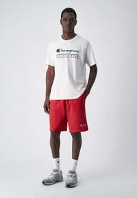 Biała koszulka polo z krótkim rękawem z logo "Champion ATHLETICWEAR", w połączeniu z czerwonymi szortami z małym logo. Uzupełnione szarymi sneakerami i białymi skarpetkami.