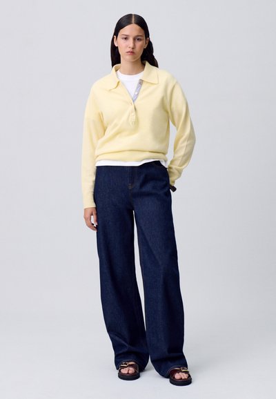 Suéter amarillo con cuello y botones, superpuesto a una camisa blanca, combinado con jeans de mezclilla oscuros de pierna ancha y sandalias negras con un acento dorado.