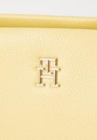 Borsa in pelle gialla con superficie testurizzata e un logo metallico in un design geometrico. La minuteria è tonalità oro e lucida.