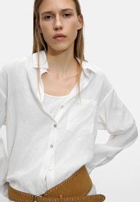 Biała, długosleeve koszula z lekkiego materiału z kieszenią, zapinana na guziki, nałożona na jasny top bez rękawów, zestawiona z brązowym paskiem z ćwiekami.