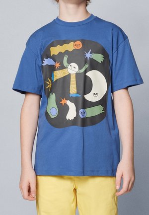 Blå t-shirt med abstrakte farverige former, smilende måne og legesyge ansigter på en sort firkantet baggrund båret af et barn i gule bukser.
