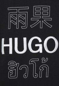 Schwarzer Stoff mit weißen umrandeten chinesischen und thailändischen Schriftzeichen oberhalb und unterhalb des fetten, weißen Sticktexts "HUGO".