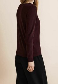 Woman wearing a long-sleeve burgundy sweater and black pants, standing sideways against a plain light background.