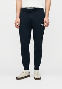 Άνδρας που φοράει μαύρο παντελόνι jogger με κορδόνι, λευκά αθλητικά παπούτσια και λευκές κάλτσες, στεκόμενος μπροστά σε απλό φόντο.