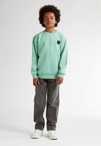 Sudadera verde menta con puños y dobladillo acanalados, con un pequeño parche de logo negro. Combinada con pantalones de pana grises y zapatillas blancas.