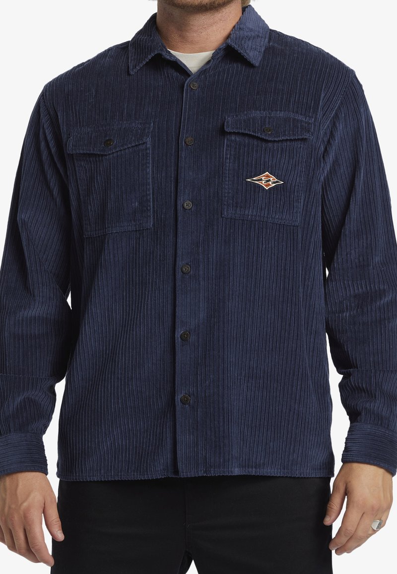 Navy corduroy shirt with long sleeves, two chest pockets, and a small embroidered logo. Features vertical ribbing and classic button closure.
