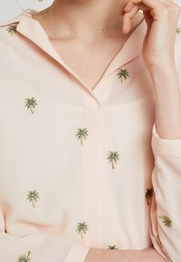 Jasnoróżowa bluzka z haftowanymi zielonymi i złotymi palmami, częściowo widoczny kołnierzyk oraz osoba dotykająca swojej twarzy.